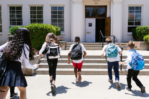 Young students run towards the school doors with their backpacks on representing the back-to-school season and part-time job opportunities.