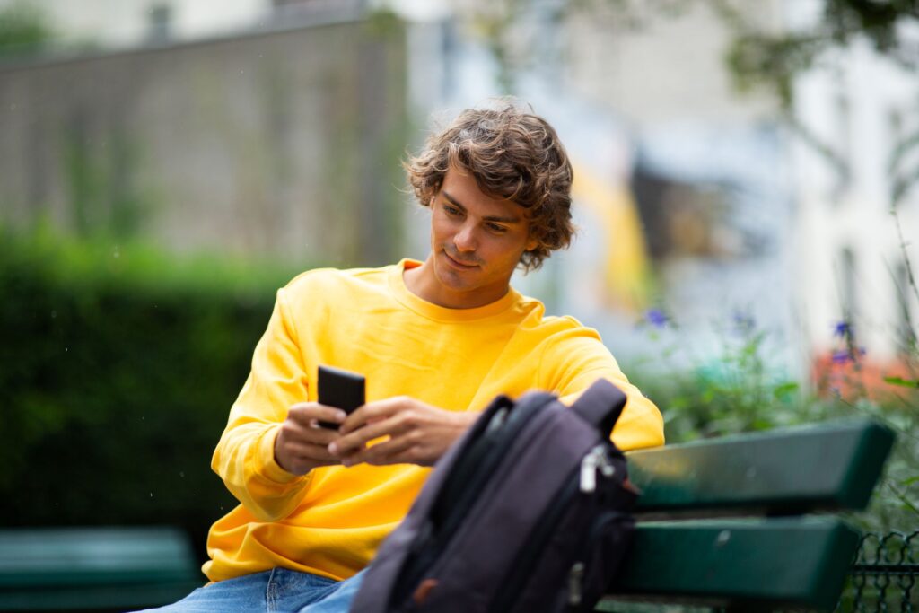 A young adult sits on a park bench in Riverside Park, La Crosse, using a smartphone to enhance their online presence for job hunting.